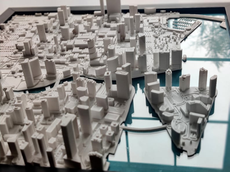 Puede incluir: Un modelo 3D blanco de un horizonte de ciudad, con edificios y carreteras, colocado en un marco de vidrio con un fondo azul.