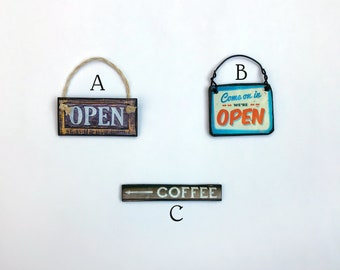 May include: Three wooden signs with different messages. The first sign says "OPEN" in white letters on a brown background. The second sign says "Come on in we're OPEN" in orange letters on a blue background. The third sign says "COFFEE" in white letters on a brown background with an arrow pointing to the left.