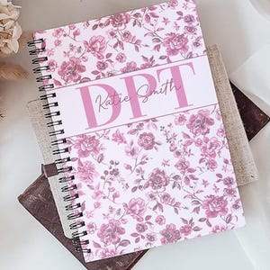 May include: A spiral-bound notebook featuring a pink and white floral pattern with a central horizontal band containing the letters DPT and the name Katie Smith in a script font, positioned alongside a gold pen and a glass mug of coffee.