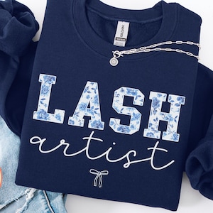 Peut inclure: Un sweat-shirt bleu marine à col rond avec le texte LASH en lettres capitales à motif floral bleu et blanc, suivi du mot artist en écriture cursive blanche et d'une petite icône de nœud en ligne blanche centrée sous le texte.