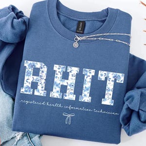 May include: An indigo blue crewneck sweatshirt featuring the white text RHIT in a block font filled with a blue floral pattern. Below the letters, the text reads registered health information technician with a small white bow icon underneath.