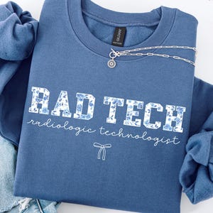 Peut inclure: Un sweat-shirt bleu avec le texte "RAD TECH" en lettres blanches avec un motif floral bleu. Le texte "radiologic technologist" est écrit en blanc sous le texte principal.