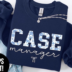 May include: Navy blue sweatshirt with the words "CASE manager" in white block letters filled with a blue floral pattern. A silver necklace with a pendant is draped across the front. The words "SUPER SOFT" are in a black oval.