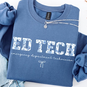 May include: A blue sweatshirt with the text "ED TECH emergency department technician" in white letters with a floral pattern. The text is surrounded by a white border.