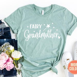 K&ouml;nnte beinhalten: Ein heather vintage mintfarbenes T-Shirt mit wei&szlig;em Schriftzug Fairy Grandmother, erg&auml;nzt durch eine Grafik eines Zauberstabs und Sternen. Das T-Shirt ist kombiniert mit einer dunkelblauen Jeans, einem Strau&szlig; rosa und wei&szlig;er Blumen und wei&szlig;en Turnschuhen.