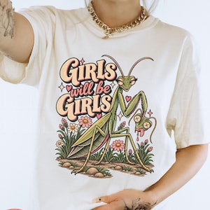 May include: A cream-coloured t-shirt featuring a graphic print of a green praying mantis holding a smaller insect head. The text "GIRLS will be GIRLS" is printed in a retro font above the insect, surrounded by small pink hearts and flowers.
