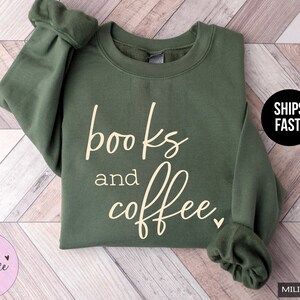 May include: Military green sweatshirt with the words "books and coffee" in a cursive font. The sweatshirt has a crew neck and long sleeves. The brand name "Stella & Grace Tees" is in a pink circle.