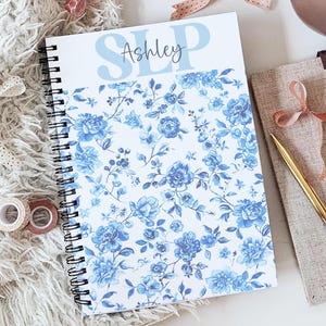 May include: A spiral-bound notebook featuring a white cover with a blue floral pattern and the text "Ashley" printed in a script font over the letters "SLP" in light blue. The notebook has a black wire binding along the left side edge.