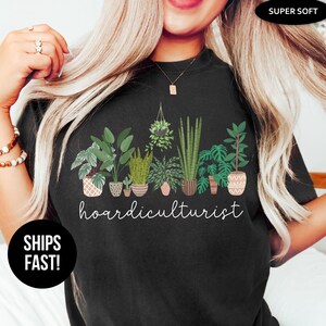 Op de afbeelding: Zwart T-shirt met een botanisch ontwerp met diverse potplanten en het woord "hoardiculturist" in witte letters. Het shirt is gelabeld met "Super Soft" en heeft een ronde hals. De merknaam "Stella & Grace Tees" is ook zichtbaar.