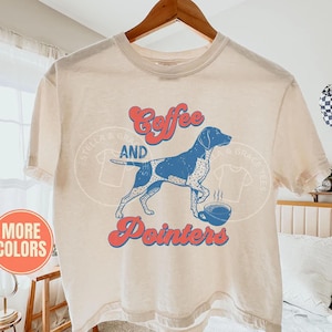 May include: An ivory short-sleeve t-shirt featuring a blue graphic of a pointer dog standing next to a steaming cup of coffee. The text "Coffee" and "Pointers" is printed in a red retro-style font above and below the dog illustration.