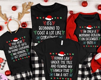May include: Black Christmas-themed t-shirts with festive text and Santa hats. The shirts feature phrases like "It's beginning to cost a lot like Christmas" and "I'm only a morning person on December 25th." The shirts are part of a holiday collection.