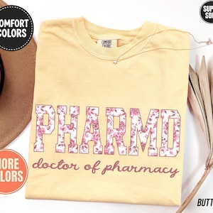 May include: A butter yellow short-sleeve t-shirt featuring the text PHARMD in a block font filled with a pink floral pattern, with the words doctor of pharmacy printed below in a cursive script. The shirt has a Comfort Colors brand label.