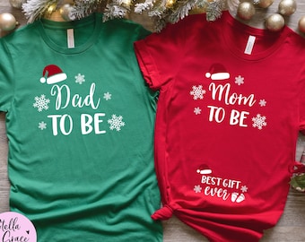 May include: Two Christmas-themed t-shirts, one green and one red. The green shirt says "Dad TO BE" with a Santa hat and snowflakes. The red shirt says "Mom TO BE" with a Santa hat and snowflakes, and "Best Gift Ever".