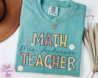 Può includere: Una maglietta turchese Comfort Colors con le parole "MATH Mrs. Anderson TEACHER" in un font retrò colorato. La maglietta presenta grafiche di margherite e stelle. Una collana d'oro con un ciondolo è drappeggiata sulla maglietta.
