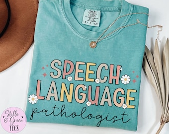 May include: A turquoise Comfort Colors t-shirt with the words "SPEECH LANGUAGE pathologist" in a colorful, retro font. The shirt features daisy accents and a gold necklace. The Stella & Grace Tees logo is in the lower left corner.