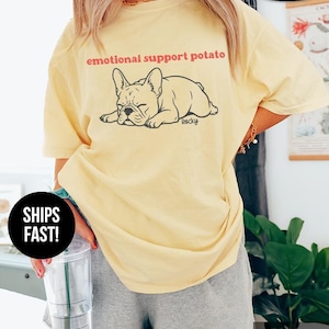 May include: A light yellow t-shirt with the text "emotional support potato" above a black illustration of a sleeping French bulldog. The shirt is labeled "Banana" at the bottom right.