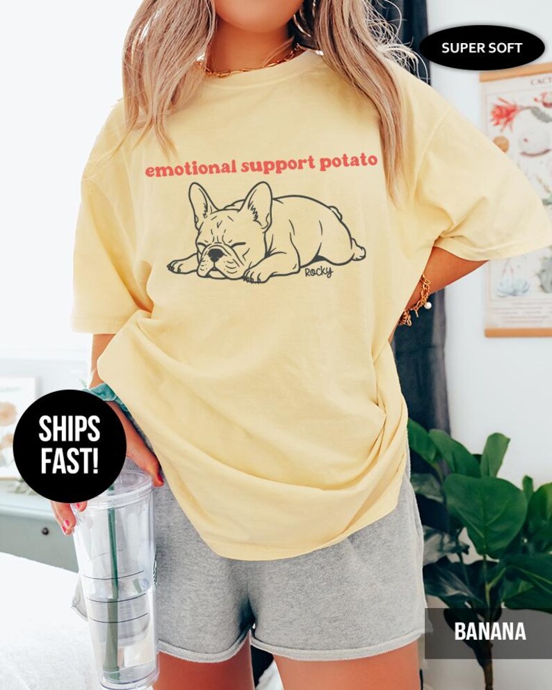 May include: A light yellow t-shirt with the text "emotional support potato" above a black illustration of a sleeping French bulldog. The shirt is labeled "Banana" at the bottom right.