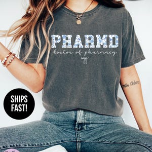 Puede incluir: Una camiseta gris con un gr&aacute;fico blanco que dice "PHARMD doctor of pharmacy" con un patr&oacute;n floral azul. La camiseta es s&uacute;per suave.