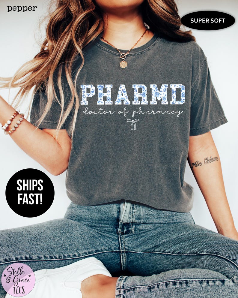 Puede incluir: Una camiseta gris con un gr&aacute;fico blanco que dice "PHARMD doctor of pharmacy" con un patr&oacute;n floral azul. La camiseta es s&uacute;per suave.