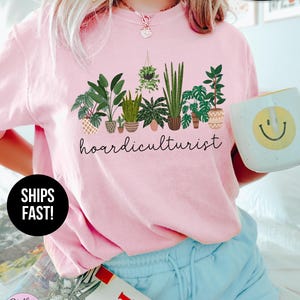 Op de afbeelding: Lichtroze T-shirt met het woord "hoardiculturist" in zwart schrift onder een rij potplanten. Het shirt heeft de woorden "SUPER SOFT" en "SHIPS FAST!" erop gedrukt. Een mok met een gele smiley wordt in de hand gehouden.