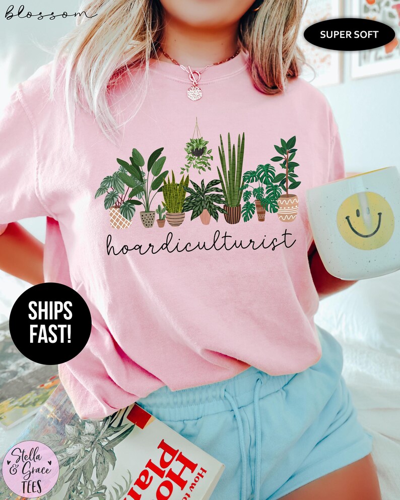 Op de afbeelding: Lichtroze T-shirt met het woord "hoardiculturist" in zwart schrift onder een rij potplanten. Het shirt heeft de woorden "SUPER SOFT" en "SHIPS FAST!" erop gedrukt. Een mok met een gele smiley wordt in de hand gehouden.