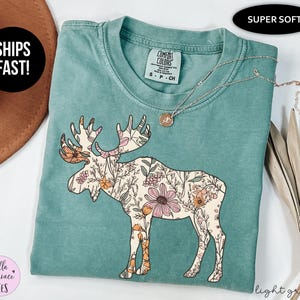 May include: A sage green short-sleeve t-shirt featuring a graphic of a moose filled with a floral pattern in shades of pink, orange, and cream. The shirt has a crew neckline and a Comfort Colors brand label on the inside collar, size small.