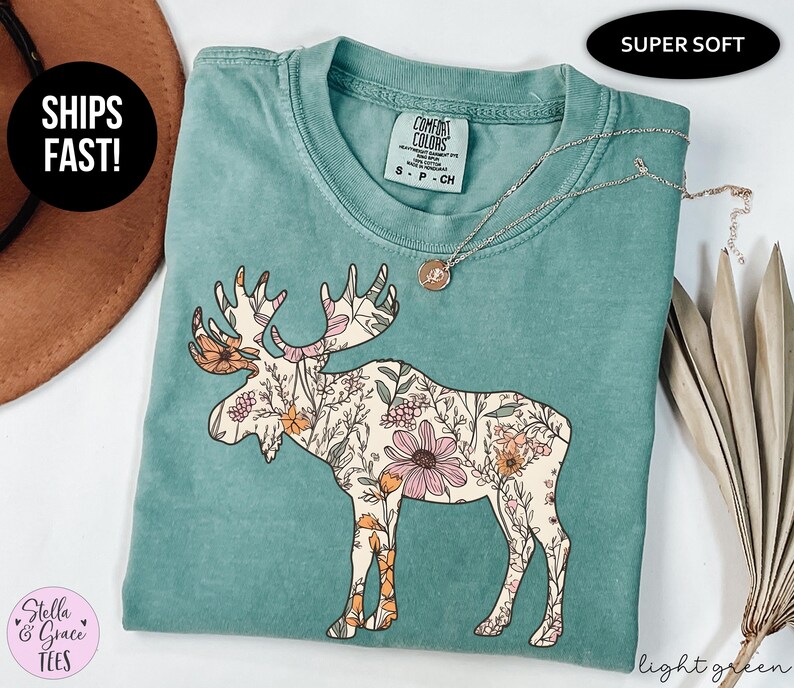 May include: A sage green short-sleeve t-shirt featuring a graphic of a moose filled with a floral pattern in shades of pink, orange, and cream. The shirt has a crew neckline and a Comfort Colors brand label on the inside collar, size small.