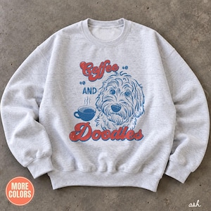 May include: A light gray crewneck sweatshirt featuring a graphic print of a doodle dog and a coffee cup. The text reads Coffee AND Doodles in red and blue lettering. The design includes small coffee bean icons and steam rising from the cup.