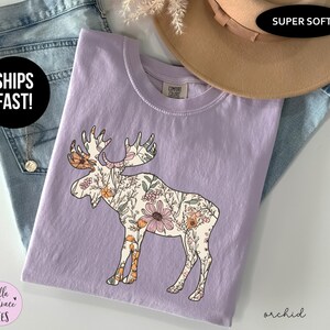 May include: A light purple short-sleeved t-shirt featuring a graphic of a moose filled with a floral pattern in shades of orange, pink, and green. The shirt has a crew neckline and a Comfort Colors brand label on the inside collar area.