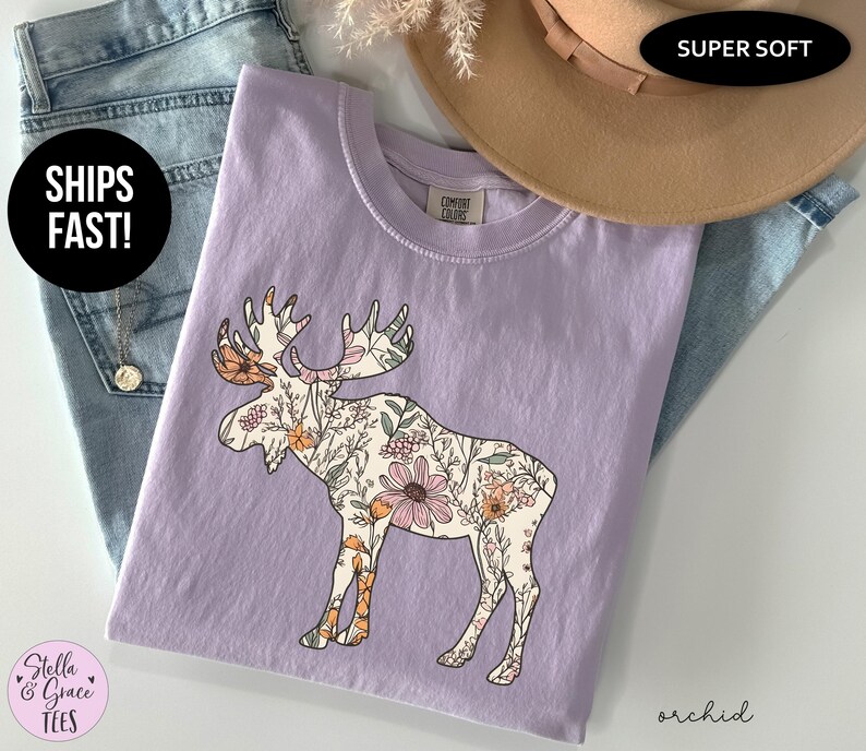 May include: A light purple short-sleeved t-shirt featuring a graphic of a moose filled with a floral pattern in shades of orange, pink, and green. The shirt has a crew neckline and a Comfort Colors brand label on the inside collar area.