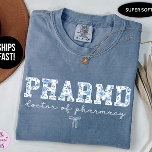 Puede incluir: Una camiseta azul con un dise&ntilde;o floral blanco que dice "PHARMD doctor of pharmacy" en letras blancas. La camiseta est&aacute; hecha de una tela suave y c&oacute;moda y es perfecta para el uso diario.