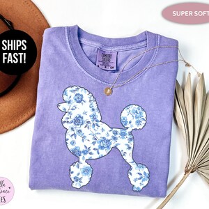 May include: A lilac-colored t-shirt featuring a floral poodle design in white and blue. The shirt has a crew neck and short sleeves. A gold necklace is draped across the shirt. The words "SUPER SOFT" are in a pink oval.