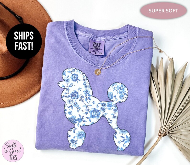 May include: A lilac-colored t-shirt featuring a floral poodle design in white and blue. The shirt has a crew neck and short sleeves. A gold necklace is draped across the shirt. The words "SUPER SOFT" are in a pink oval.