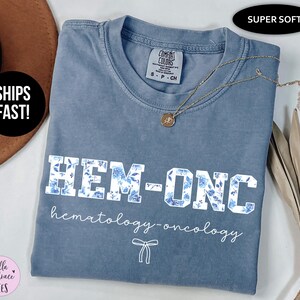 May include: A light blue t-shirt with the text "HEM-ONC hematology-oncology" in white lettering with blue floral accents. The shirt is made of a soft, comfortable fabric.