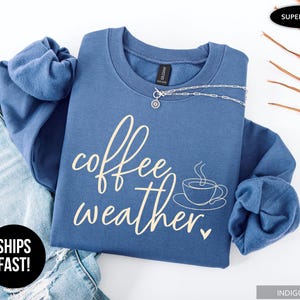May include: Indigo blue sweatshirt with the words "coffee weather" in a cursive font, along with a coffee cup graphic. A silver necklace is draped across the front. The sweatshirt is folded on top of a pair of blue jeans.