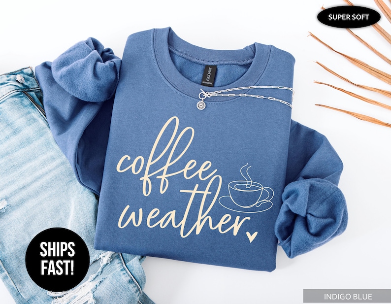 May include: Indigo blue sweatshirt with the words "coffee weather" in a cursive font, along with a coffee cup graphic. A silver necklace is draped across the front. The sweatshirt is folded on top of a pair of blue jeans.