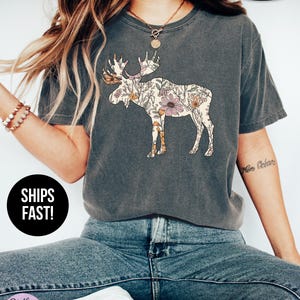 May include: A charcoal gray short-sleeve t-shirt featuring a graphic of a moose filled with a floral pattern in shades of pink, orange, and green. The shirt has a crew neckline and a relaxed fit. Text on the image includes pepper, SUPER SOFT, SHIPS FAST!, and Stella & Grace TEES.