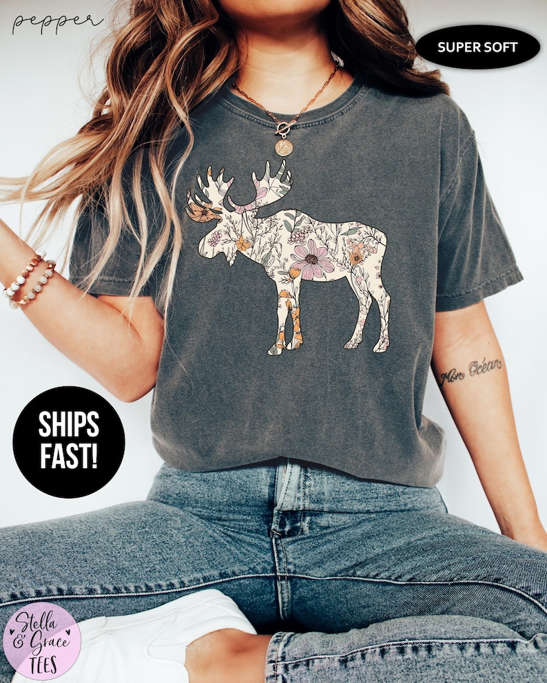 May include: A charcoal gray short-sleeve t-shirt featuring a graphic of a moose filled with a floral pattern in shades of pink, orange, and green. The shirt has a crew neckline and a relaxed fit. Text on the image includes pepper, SUPER SOFT, SHIPS FAST!, and Stella & Grace TEES.