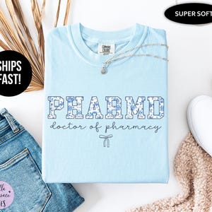 Puede incluir: Camiseta azul claro con un dise&ntilde;o de estampado floral que dice "PHARMD doctor of pharmacy" en una fuente universitaria.
