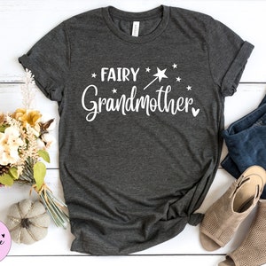 K&ouml;nnte beinhalten: Ein dunkelgraues T-Shirt mit kurzen &Auml;rmeln und dem wei&szlig;en Schriftzug "FAIRY Grandmother" auf der Vorderseite. Das Design enth&auml;lt eine kleine Grafik eines Zauberstabs mit Sternspitze, mehrere verstreute Sterne und ein kleines Herzsymbol.