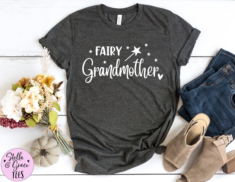 K&ouml;nnte beinhalten: Ein dunkelgraues T-Shirt mit kurzen &Auml;rmeln und dem wei&szlig;en Schriftzug "FAIRY Grandmother" auf der Vorderseite. Das Design enth&auml;lt eine kleine Grafik eines Zauberstabs mit Sternspitze, mehrere verstreute Sterne und ein kleines Herzsymbol.
