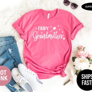K&ouml;nnte beinhalten: Ein pinkfarbenes Kurzarm-T-Shirt mit dem wei&szlig;en Schriftzug "FAIRY Grandmother", einer Grafik eines Zauberstabs mit Stern und kleinen Sternen. Das T-Shirt ist zusammen mit einer blauen Jeans, wei&szlig;en Turnschuhen und einem Blumenstrau&szlig; abgebildet.