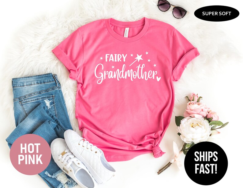 K&ouml;nnte beinhalten: Ein pinkfarbenes Kurzarm-T-Shirt mit dem wei&szlig;en Schriftzug "FAIRY Grandmother", einer Grafik eines Zauberstabs mit Stern und kleinen Sternen. Das T-Shirt ist zusammen mit einer blauen Jeans, wei&szlig;en Turnschuhen und einem Blumenstrau&szlig; abgebildet.