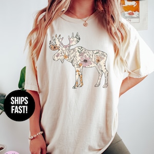 May include: An ivory short-sleeve t-shirt featuring a large graphic of a moose silhouette filled with a colorful floral pattern. The shirt has a crew neckline and a relaxed fit. Text on the image includes 'ivory', 'SUPER SOFT', and 'SHIPS FAST!'.