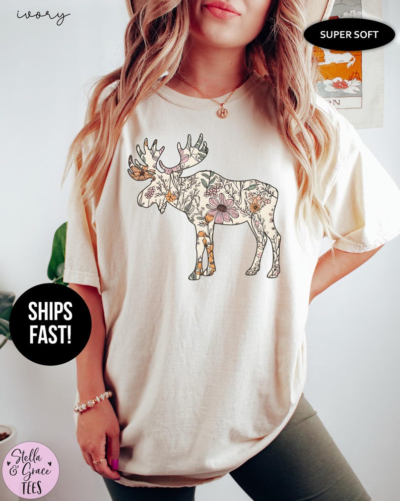 May include: An ivory short-sleeve t-shirt featuring a large graphic of a moose silhouette filled with a colorful floral pattern. The shirt has a crew neckline and a relaxed fit. Text on the image includes 'ivory', 'SUPER SOFT', and 'SHIPS FAST!'.