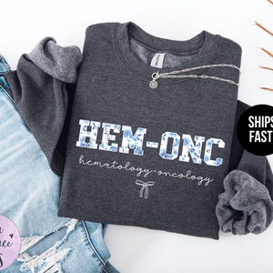 May include: Dark grey sweatshirt with the text "HEM-ONC hematology-oncology" in white with blue floral accents.