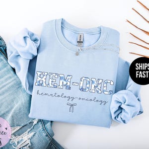 May include: Light blue sweatshirt with the text "HEM-ONC hematology-oncology" in a blue and white patterned font. A small white ribbon is below the text.