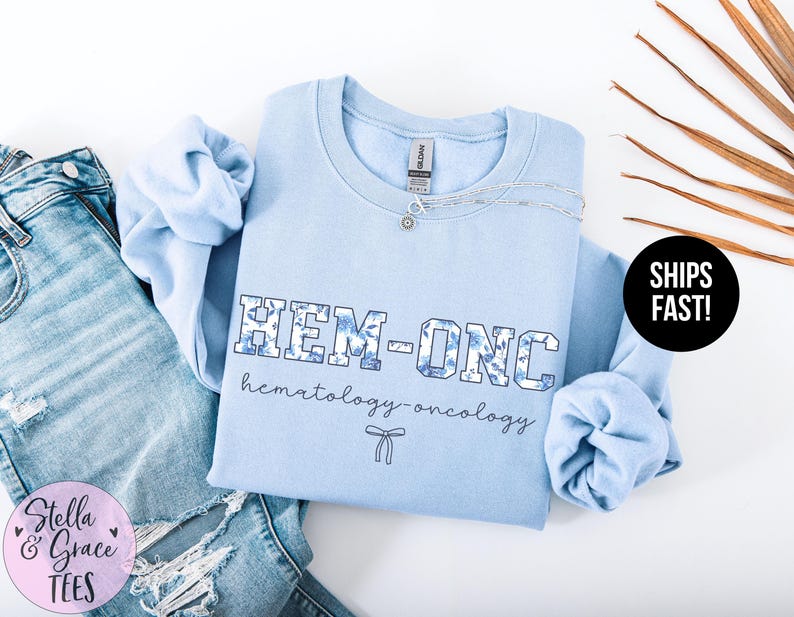 May include: Light blue sweatshirt with the text "HEM-ONC hematology-oncology" in a blue and white patterned font. A small white ribbon is below the text.