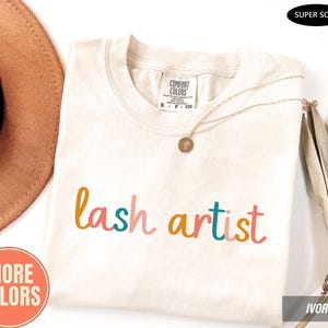 May include: An ivory short-sleeved t-shirt with the text "lash artist" printed in multicoloured script letters across the chest. The shirt features a Comfort Colors brand label at the inner neckline and a small gold-toned circular pendant necklace.