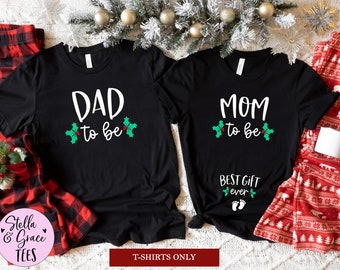 May include: Black t-shirts with white text that reads "DAD to be" and "MOM to be" with holly accents. The "MOM" shirt also says "BEST GIFT ever" with baby feet. The shirts are part of a Christmas-themed set.